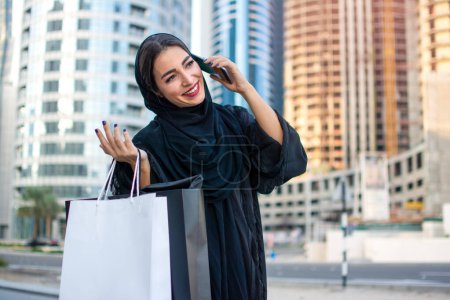 Photo for Arab woman in Abaya talking on mobile phone while holding shopping bags and walking on city street - Royalty Free Image