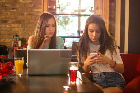 Téléchargez les photos : Adolescentes avec ordinateur portable et téléphones prenant un verre dans un restaurant - en image libre de droit