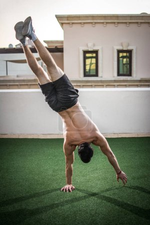 Photo for Back view of handsome shirtless man doing handstand on one hand outdoors. - Royalty Free Image