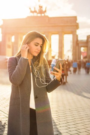 Foto de Hermosa joven escuchando música en el teléfono móvil mientras está de pie cerca de la puerta de Brandenburgo en Berlín. - Imagen libre de derechos