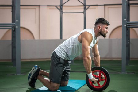 Téléchargez les photos : Vue latérale du bel homme travaillant avec une roue de fitness dans la salle de gym. - en image libre de droit