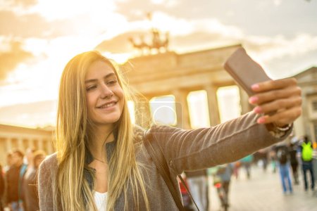 Foto de Chica sonriente con teléfono tomar foto selfie frente a la puerta de Brandenburgo en Berlín. - Imagen libre de derechos