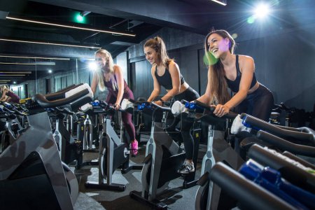 Téléchargez les photos : Joyeuses femmes sportives chevauchant des vélos d'exercice en cours de cyclisme dans la salle de gym. - en image libre de droit