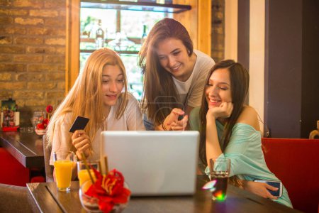 Téléchargez les photos : Trois adolescentes faisant du shopping en ligne ensemble dans un café - en image libre de droit