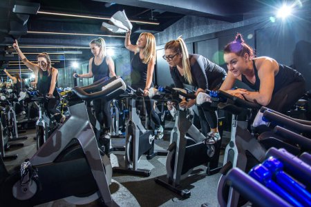 Photo for Group of cheerful sporty girls training on fitness bikes and having fun together on cycling class in gym. - Royalty Free Image