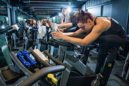 Photo for Group of young women in sportswear stretching their legs on exercise bikes before cycling class in gym. - Royalty Free Image