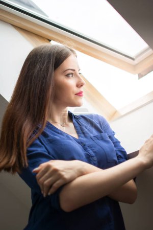 Téléchargez les photos : Jeune femme regardant par la fenêtre du grenier - en image libre de droit