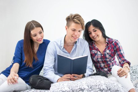 Téléchargez les photos : Trois jeunes amis lisant ensemble un livre alors qu'ils étaient assis sur l'entraîneur à la maison - en image libre de droit