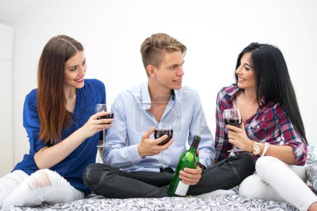 Téléchargez les photos : Trois jeunes amis assis sur le lit et buvant du vin rouge ensemble - en image libre de droit