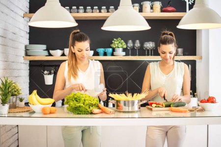 Téléchargez les photos : Sœurs jumelles cuisiner et s'amuser dans la cuisine. Ils parlent et rient. couper des légumes sur une planche à découper. - en image libre de droit