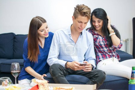 Téléchargez les photos : Trois jeunes amis utilisant un smartphone à la maison - en image libre de droit