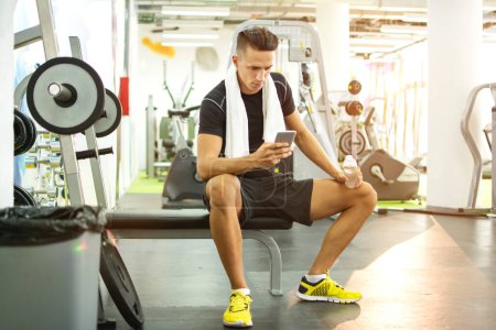 Foto de Joven hombre guapo usando el teléfono mientras hace ejercicio en el gimnasio
. - Imagen libre de derechos