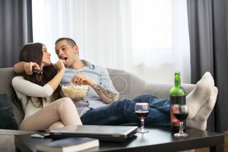Photo for Young couple eating popcorn together while sitting on sofa and watching tv - Royalty Free Image