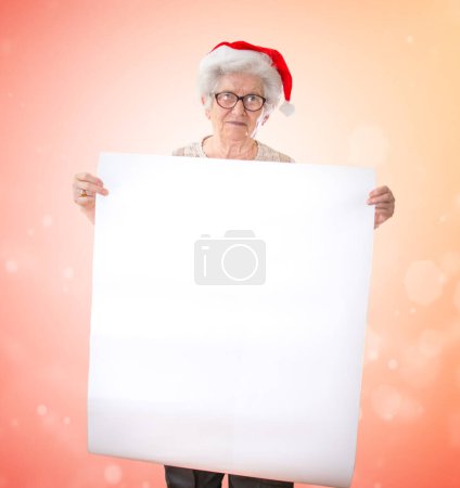 Photo for Senior woman wearing Santas hat and winter jumper holding white blank board - Royalty Free Image