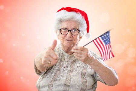 Téléchargez les photos : Femme âgée portant le chapeau de Santas montrant pouce vers le haut et tenant le drapeau américain contre fond rouge de Noël - en image libre de droit