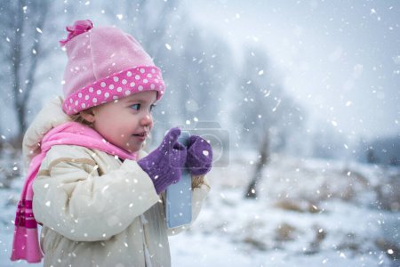 Téléchargez les photos : Petite fille avec téléphone intelligent dans le parc d'hiver pendant la journée enneigée - en image libre de droit