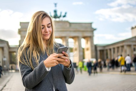 Téléchargez les photos : Belle jeune fille blonde utilisant le téléphone devant la porte de Brandebourg à Berlin, Allemagne - en image libre de droit