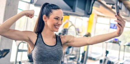 Foto de Hermosa mujer en forma flexión y mostrando sus músculos del brazo mientras toma una foto selfie en el gimnasio - Imagen libre de derechos
