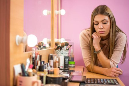 Téléchargez les photos : Belle jeune femme regardant un bureau plein de cosmétiques et choisissant ce qu'il faut appliquer sur son visage. - en image libre de droit