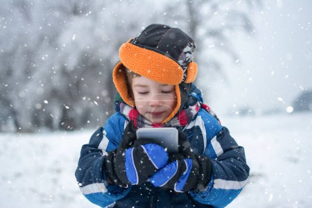 Téléchargez les photos : Portrait de petit garçon avec téléphone portable sur une journée enneigée dans un parc d'hiver - en image libre de droit