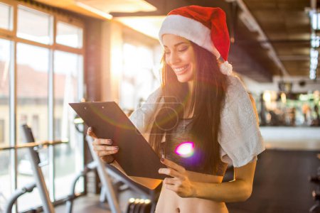 Téléchargez les photos : Entraîneur personnel féminin avec chapeau Santa tenant presse-papiers à la salle de gym. Nouvel An et concept de Noël. - en image libre de droit