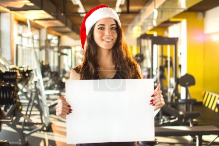 Téléchargez les photos : Belle femme de remise en forme dans le chapeau Santa tenant conseil vide. Nouvel An. Noël, vacances, fitness et concept de gym. - en image libre de droit