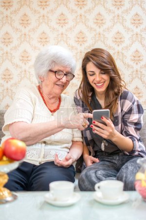 Téléchargez les photos : Grand-mère montrant quelque chose sur le téléphone intelligent à sa petite-fille à la maison - en image libre de droit
