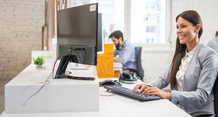 Foto de Atractiva joven trabajadora empleada de traje usando computadora mientras está sentada en su escritorio en la oficina. - Imagen libre de derechos