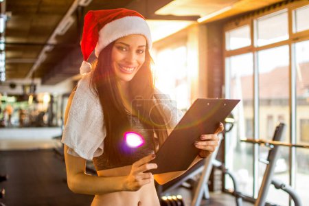 Téléchargez les photos : Entraîneur personnel féminin avec chapeau Santa tenant presse-papiers à la salle de gym. Nouvel An et concept de Noël. - en image libre de droit