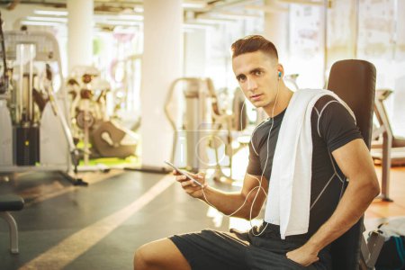 Téléchargez les photos : Beau jeune homme avec écouteurs utilisant un téléphone intelligent dans la salle de gym. - en image libre de droit