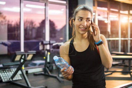 Sporty woman talking on mobile phone and holding water bottle in gym.