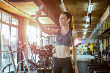 Photo for Beautiful young fit woman taking selfie photo using her smartphone after sports training in gym - Royalty Free Image