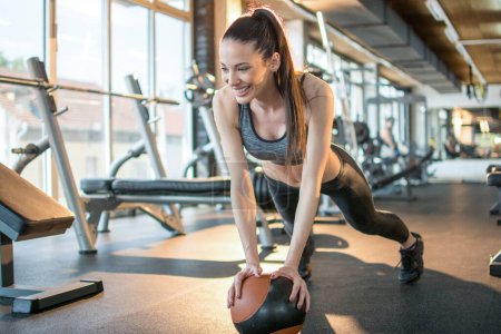 Téléchargez les photos : Souriant jeune femme en forme en vêtements de sport faire de l'exercice de planche avec balle dans la salle de gym. - en image libre de droit
