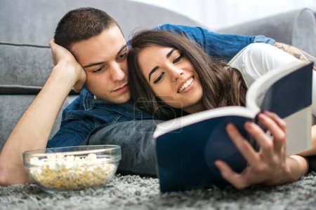 Téléchargez les photos : Jeune couple lecture livre ensemble tandis que couché sur le sol - en image libre de droit