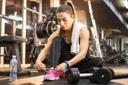 Photo for Young sporty woman tying shoes while sitting on floor in gym - Royalty Free Image
