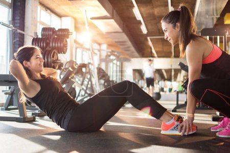 Photo for Young woman exercising sit-ups with assistance of female friend in gym. - Royalty Free Image