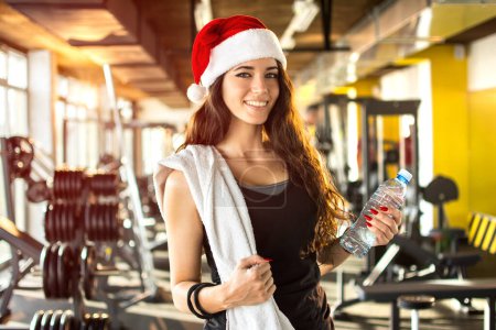 Téléchargez les photos : Jeune femme avec chapeau de Père Noël, bouteille d'eau et serviette dans la salle de gym. - en image libre de droit