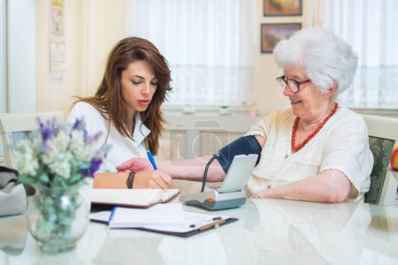 Téléchargez les photos : Jeune infirmière mesurant la pression artérielle de la femme âgée à la maison - en image libre de droit