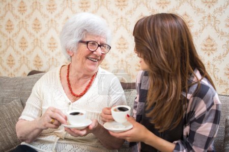 Foto de Alegre abuela y nieta con tazas de café hablando en casa - Imagen libre de derechos