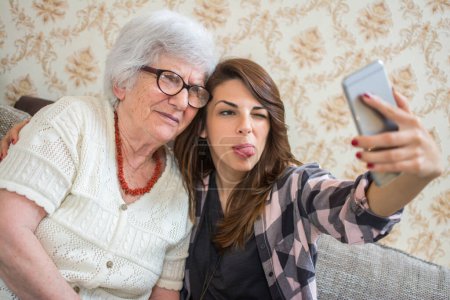 Téléchargez les photos : Petite-fille et sa grand-mère prenant selfie avec téléphone intelligent à la maison - en image libre de droit