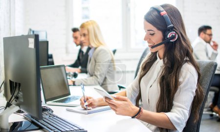 Photo for Friendly customer support female operator with headphones writing notes to notebook taken from smartphone at office - Royalty Free Image