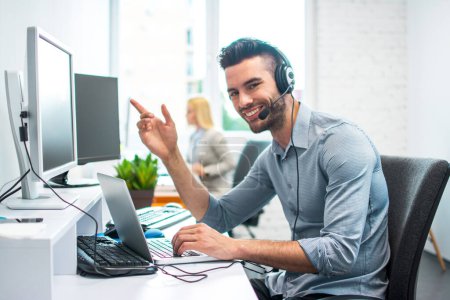 Foto de Atractivo hombre de negocios con auriculares usando computadora portátil y computadora en la oficina - Imagen libre de derechos