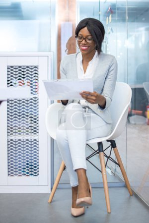 Foto de Joven mujer de negocios afroamericana sonriente o secretaria sentada en la silla de la oficina y leyendo un documento. - Imagen libre de derechos