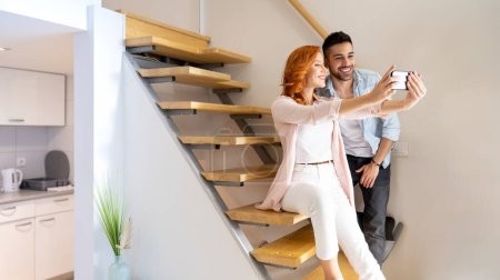 Téléchargez les photos : Mignon, jeune et heureux couple prendre un selfie ensemble dans les escaliers à la maison - en image libre de droit