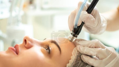 Close up of therapists hands in protective gloves holding hydrafacial tool and performing anti aging progress to beautiful young woman forehead at beauty salon.