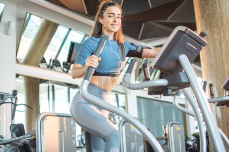 Téléchargez les photos : S'adapter jeune femme faisant de l'exercice sur une machine elliptique d'entraînement cardio au gymnase - en image libre de droit