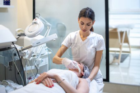 Téléchargez les photos : Esthéticienne appliquant un traitement avec un appareil moderne sur la peau des jeunes femmes au salon de spa - en image libre de droit
