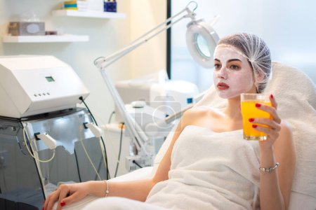 Photo for Young beautiful woman with face mask drinking orange juice at beauty salon. - Royalty Free Image