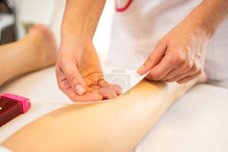 Téléchargez les photos : Gros plan d'une esthéticienne épilation jambe de femme avec bande de cire dans un spa de beauté. - en image libre de droit