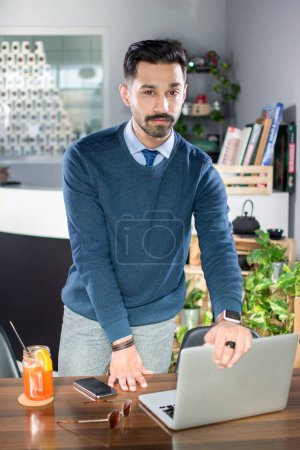 Téléchargez les photos : Portrait de beau jeune homme en tenue décontractée intelligente avec les mains sur la table de travail et ordinateur portable dans un bureau - en image libre de droit